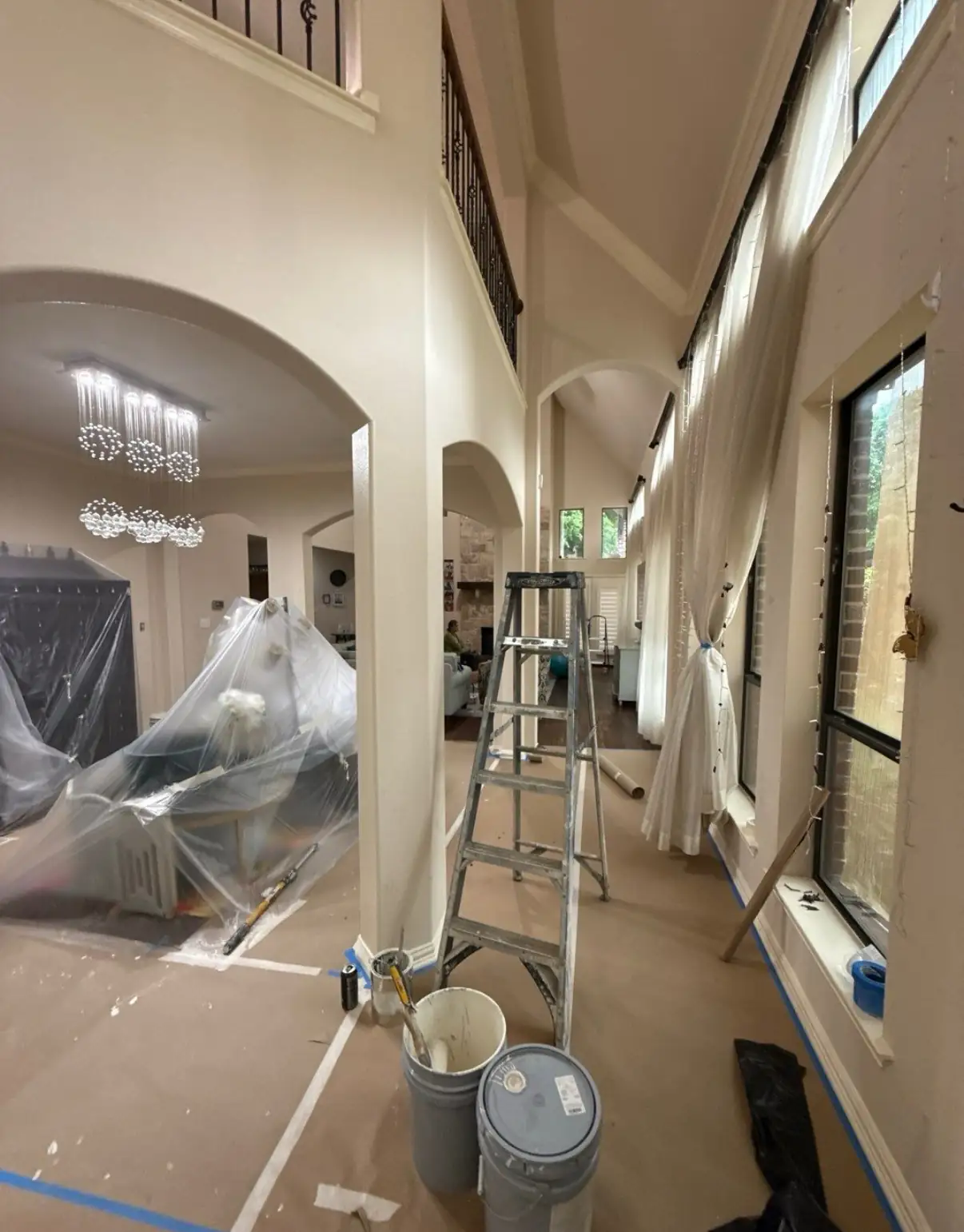 Tall great room interior prepped for painting with ladders, floor covering, and furniture wrapped in plastic