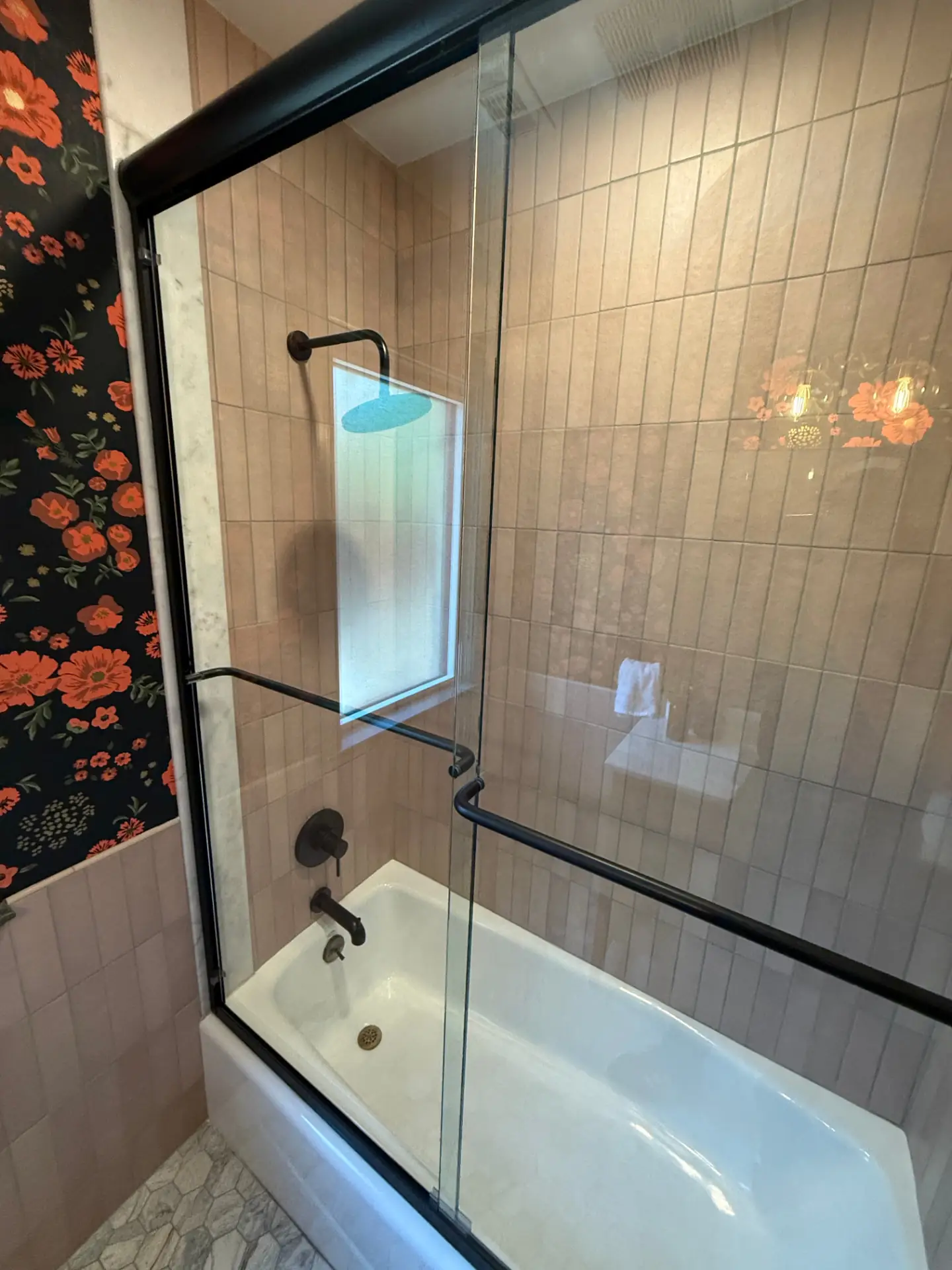 Corner view of the tub shower with black fixtures, vertical tile, and a bold floral wallpaper accent wall